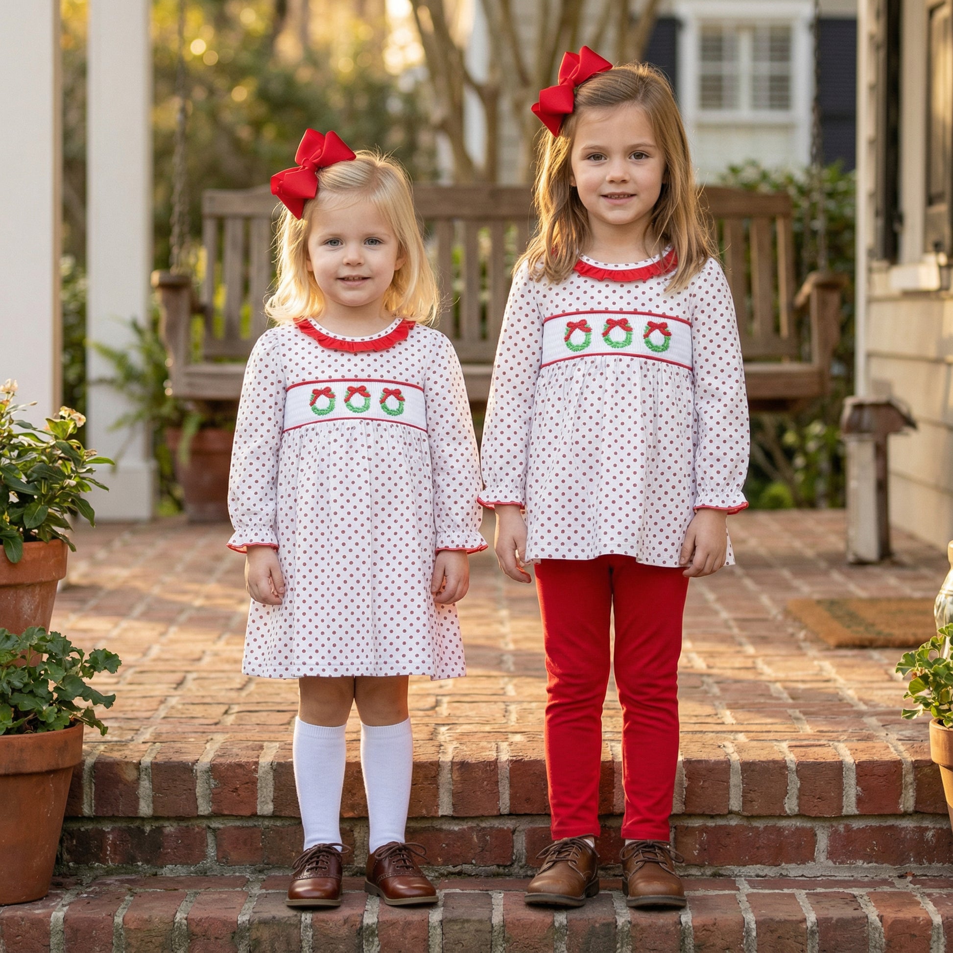 Two young girls stand side-by-side on brick steps wearing matching holiday outfits. Both girls wear white long-sleeved garments with red polka dots and smocking across the chest featuring embroidered Christmas wreaths, along with large red hair bows. The younger girl on the left wears a dress with white knee-high socks, while the older girl on the right wears a tunic top paired with red leggings. A wooden porch swing and potted plants are visible in the background.