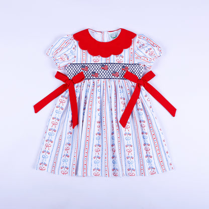 America the Beautiful hand smocked dress for girls featuring American flags, red scalloped collar, and waist bows.