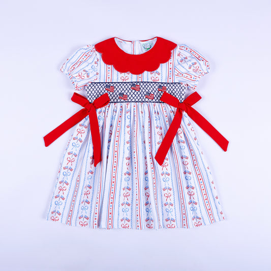 America the Beautiful hand smocked dress for girls featuring American flags, red scalloped collar, and waist bows.
