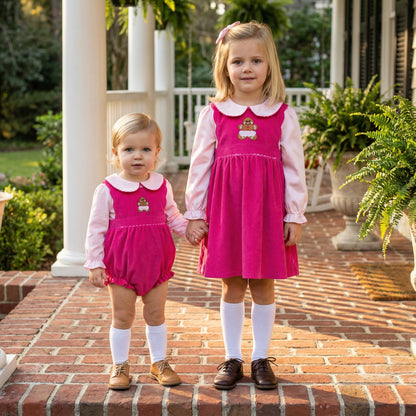 Two young girls stand on a brick porch wearing matching hot pink corduroy outfits featuring gingerbread man appliqués on the chest. The younger child wears a bubble romper over a light pink collared blouse, while the older child wears a pinafore dress over a matching shirt. Both complete the traditional look with white knee-high socks and leather dress shoes.