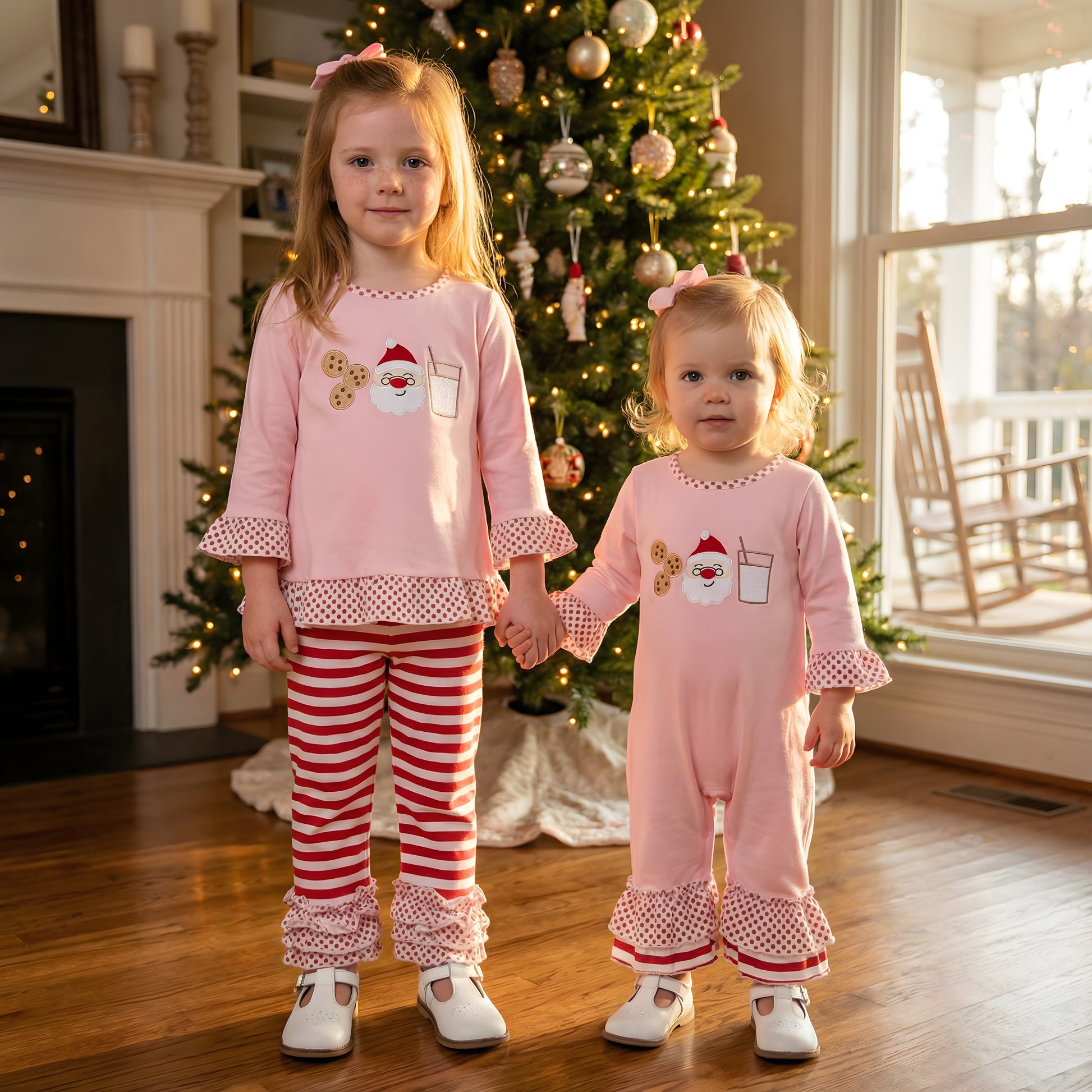 Two sisters holding hands in front of a Christmas tree wearing matching pink Girls Milk And Cookies With Santa Christmas Embroidered Pants Set and Romper.