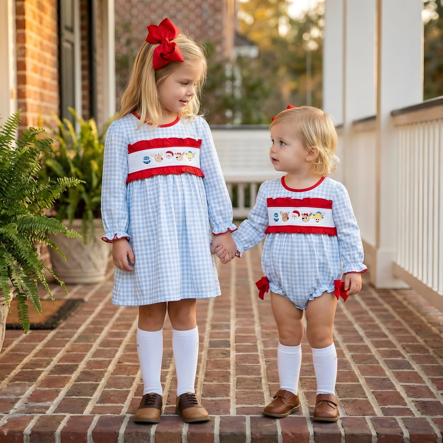 Two sisters wearing matching light blue gingham smocked Christmas outfits with red bows and white knee socks, holding hands on a brick porch. The older girl is in a smocked dress, and the younger girl is in a matching bubble romper. This image showcases toddler fashion and holiday sibling clothing.