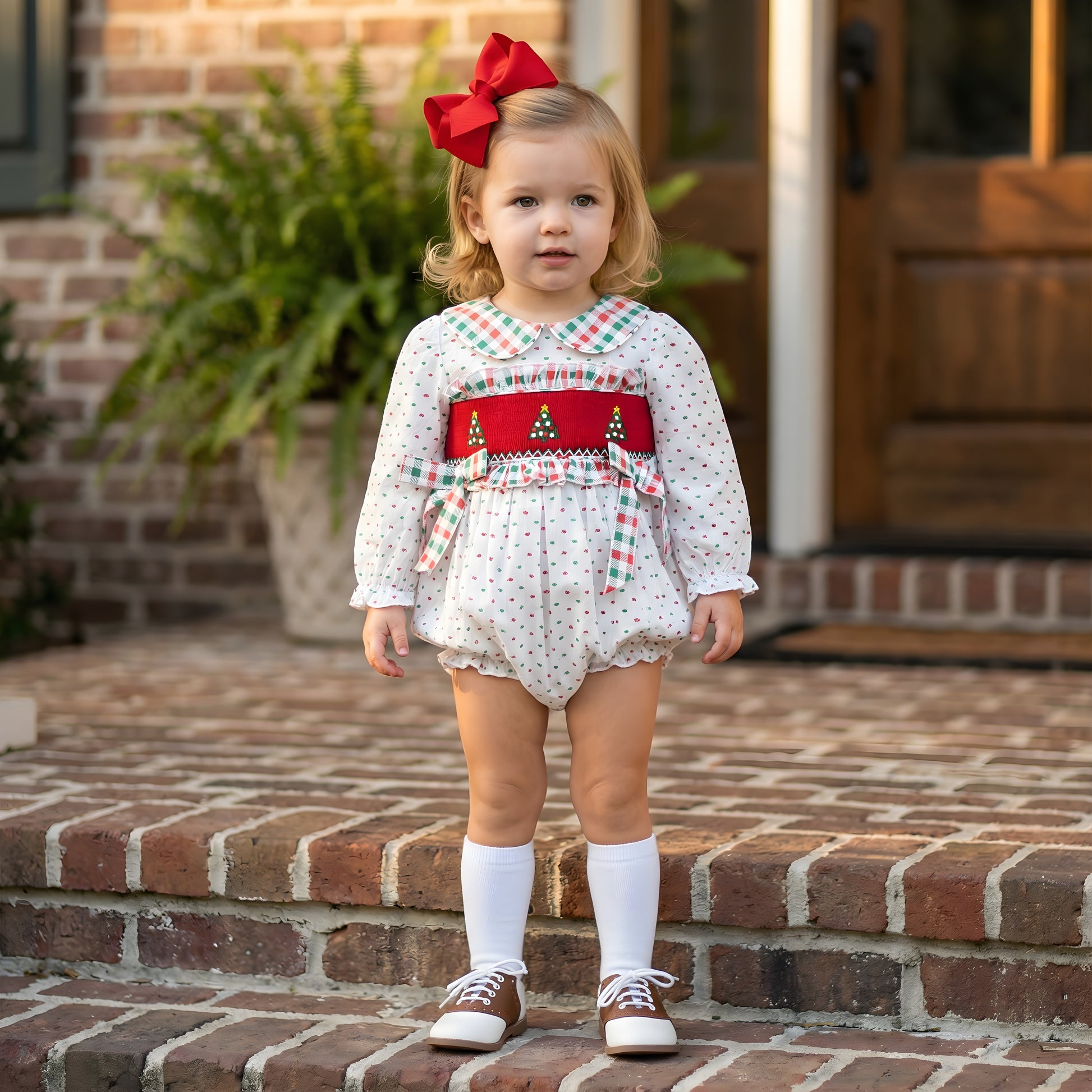 Toddler girl standing on brick steps wearing a white Girls Hand Smocked Swiss Dot Christmas Tree Bubble featuring a red smocked panel with embroidered trees, plaid trim, and a red bow for Christmas.