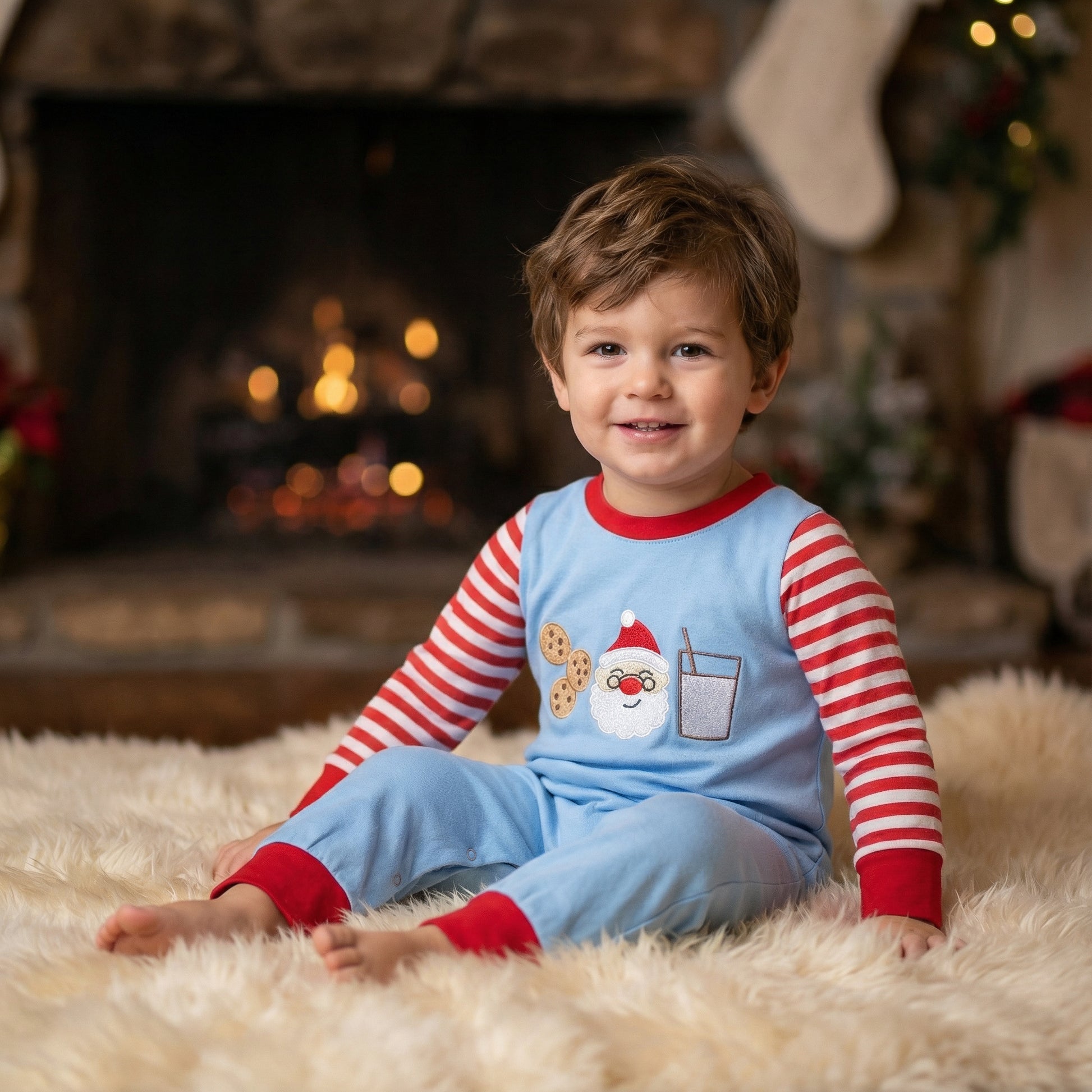 Baby boy sitting on rug by fireplace wearing blue milk and cookies Santa Christmas romper.