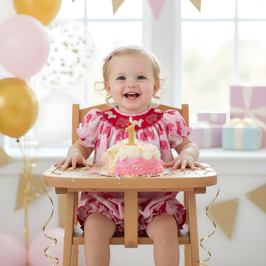Smiling baby girl sitting in a high chair celebrating her first birthday with a cake, wearing a pink sprinkle elephant bubble romper with puff sleeves.