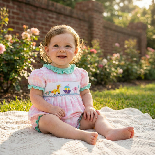 Smiling baby girl in a pink gingham hand-smocked Noah's Ark bubble romper, sitting on a blanket outdoors.