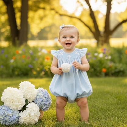 Smiling baby girl standing outdoors in a sky blue hand-smocked christening bubble romper with white crosses.