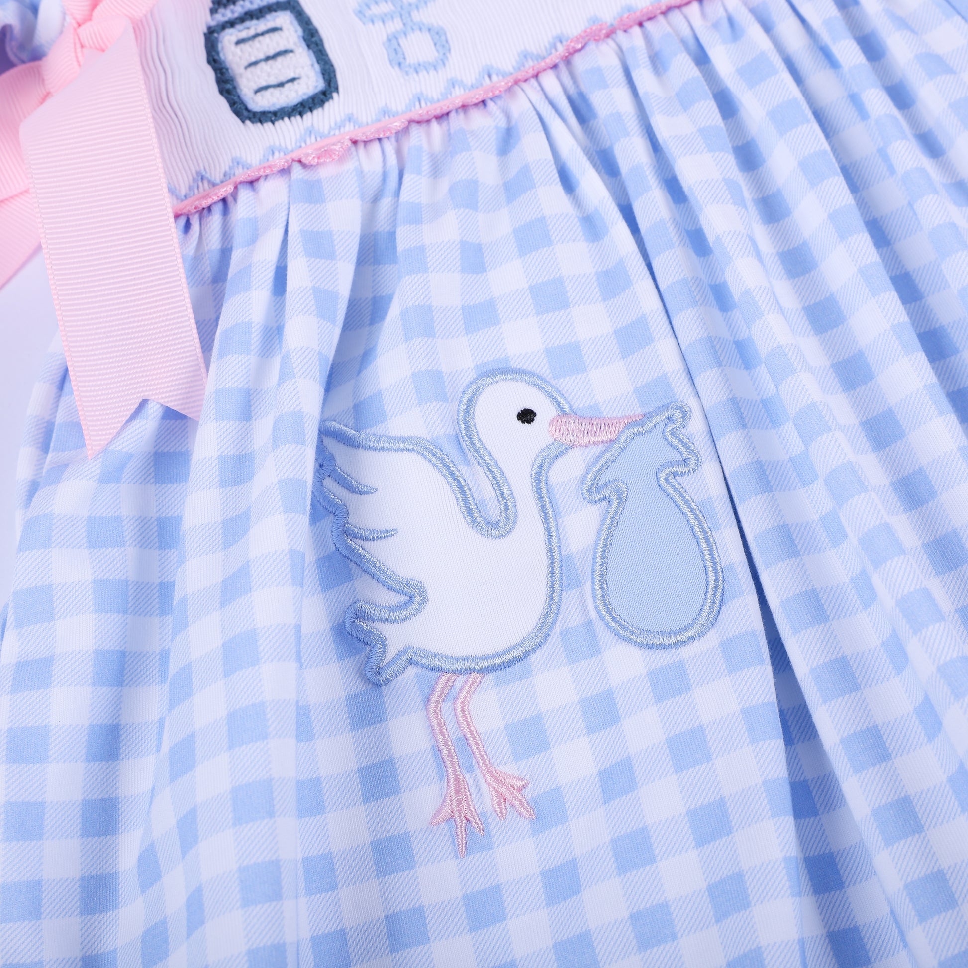 Close up of embroidered white stork delivering a blue bundle on the gathered skirt of light blue gingham baby dress.