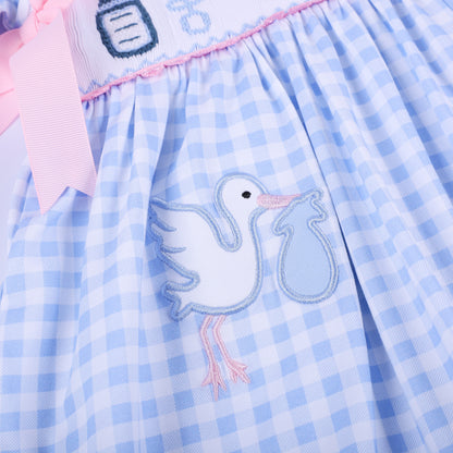 Close up of embroidered white stork delivering a blue bundle on the gathered skirt of light blue gingham baby dress.