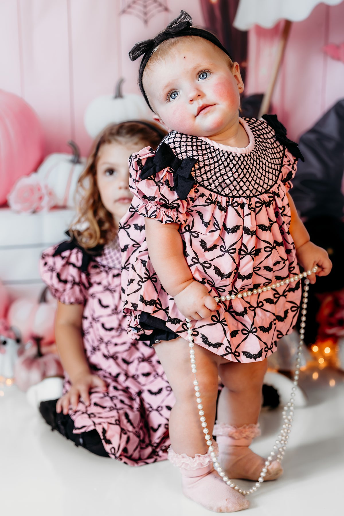 Baby girl standing in pink Batty for Bows hand smocked embroidered bats bloomer set holding pearl necklace.