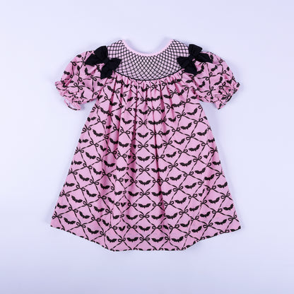 Pink hand smocked Halloween dress for girls featuring black bat bow print, black yoke embroidery, and puff sleeves.