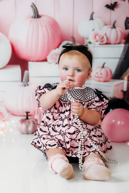 Baby girl sitting in Batty for Bows hand smocked embroidered bats bloomer set holding white pearl beads.