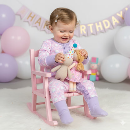 Baby sitting in a pink rocking chair wearing the Birthday Cake and Bow Girl Zippy while holding a doll.