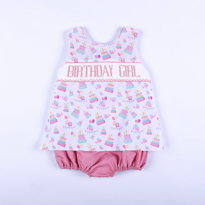 Front view of a pink birthday girl hand smocked bubble outfit with hand embroidery and cake print.