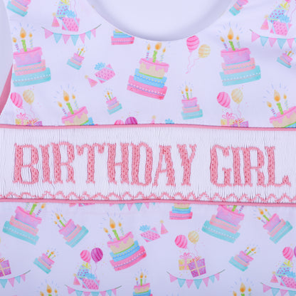 Flat lay close up of hand smocked pink birthday girl embroidery surrounded by pastel balloon and cake patterns.