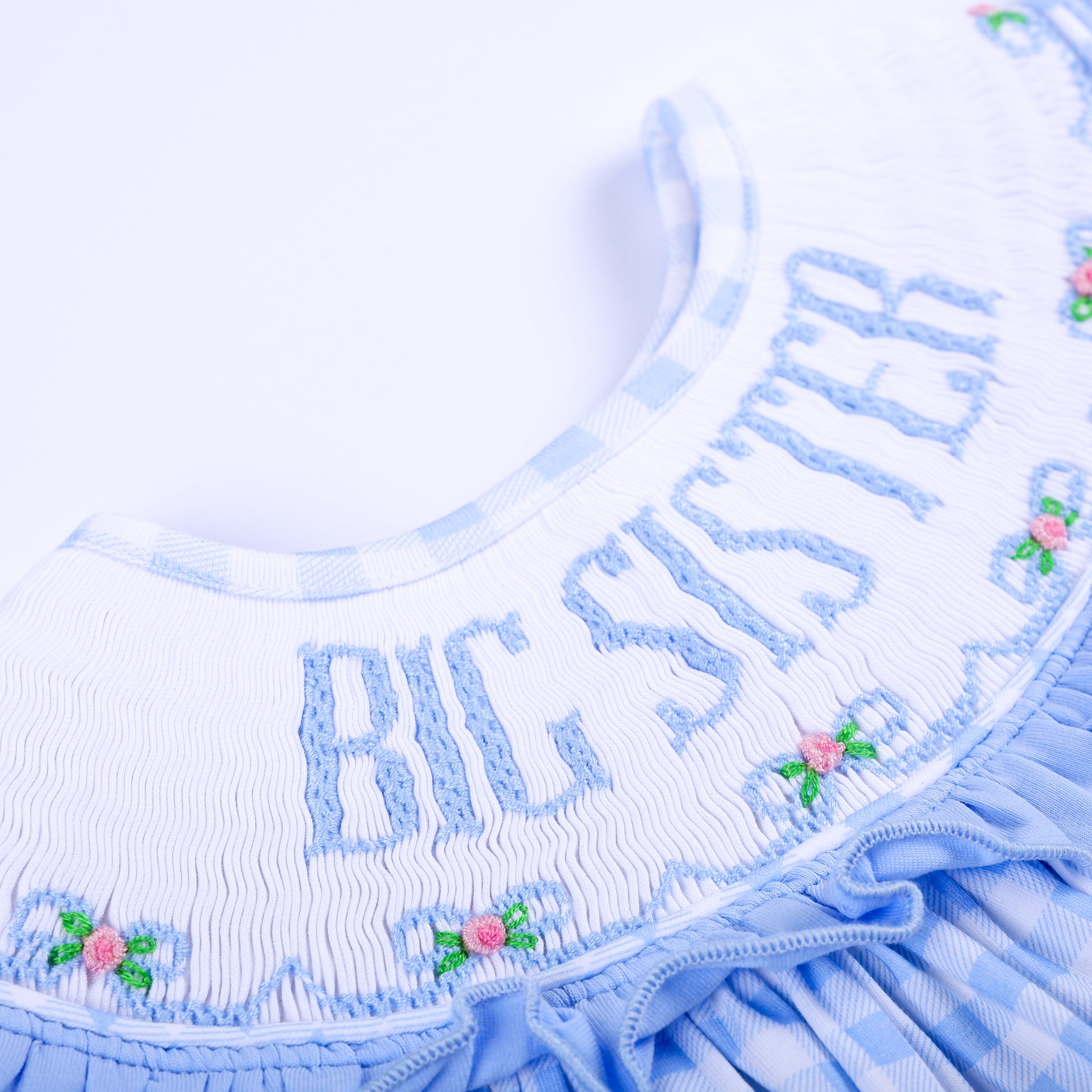 etailed close-up of hand-embroidered Big Sister lettering with pink floral bow accents on white smocking.
