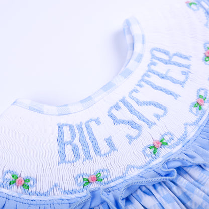 etailed close-up of hand-embroidered Big Sister lettering with pink floral bow accents on white smocking.