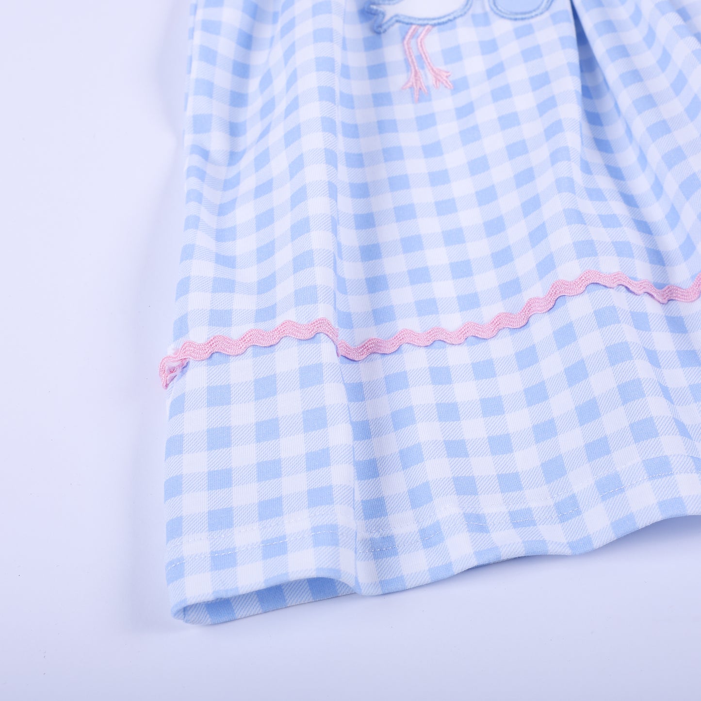 Detailed view of the pink rickrack accent trim running along the full hem of light blue gingham check smocked dress.