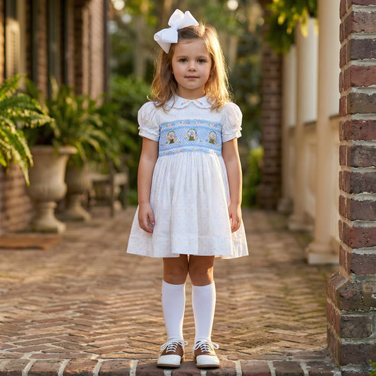 Young girl wearing a white dress with a blue hand-smocked bodice featuring embroidered flower baskets and a white bow.