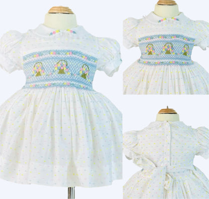 White dress with blue hand smocking, embroidered flower baskets, and pastel dot print for baby girls on a mannequin.