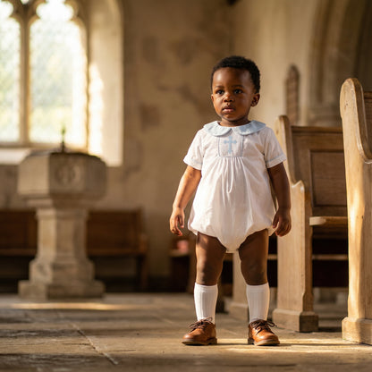 Toddler boy standing in church wearing white smocked blue cross bubble and brown shoes for christening.