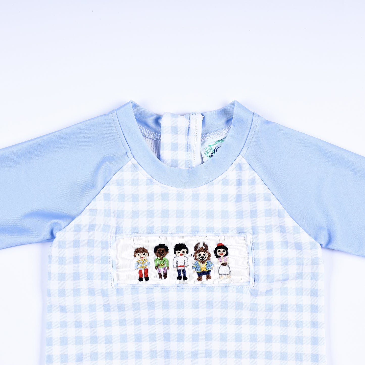 Close-up of boys hand smocked handsome prince rash guard chest featuring detailed hand embroidered fairytale figures.