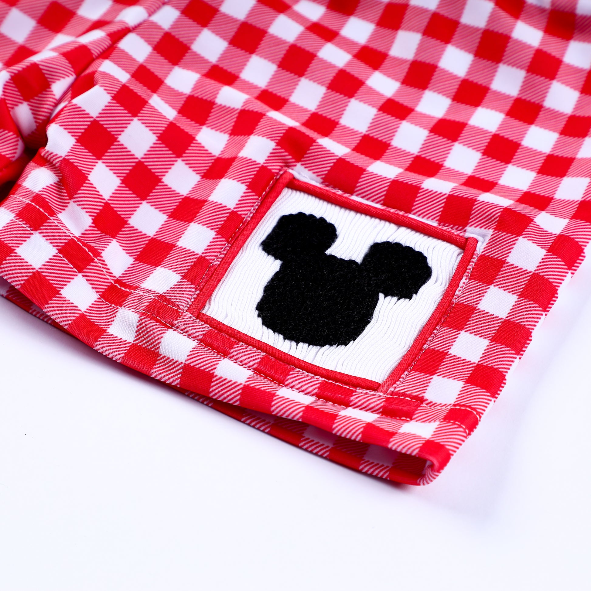 Detailed close-up of hand smocked black mouse silhouette patch on boys red and white gingham swim trunks.