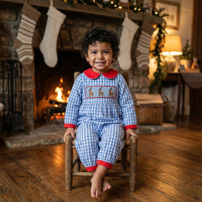 Toddler boy wearing blue gingham hand smocked reindeer romper sitting by a Christmas fireplace.