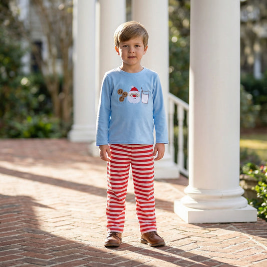 Boy standing on a brick porch wearing a light blue shirt with Santa, milk, and cookies appliqué and red and white striped pants.