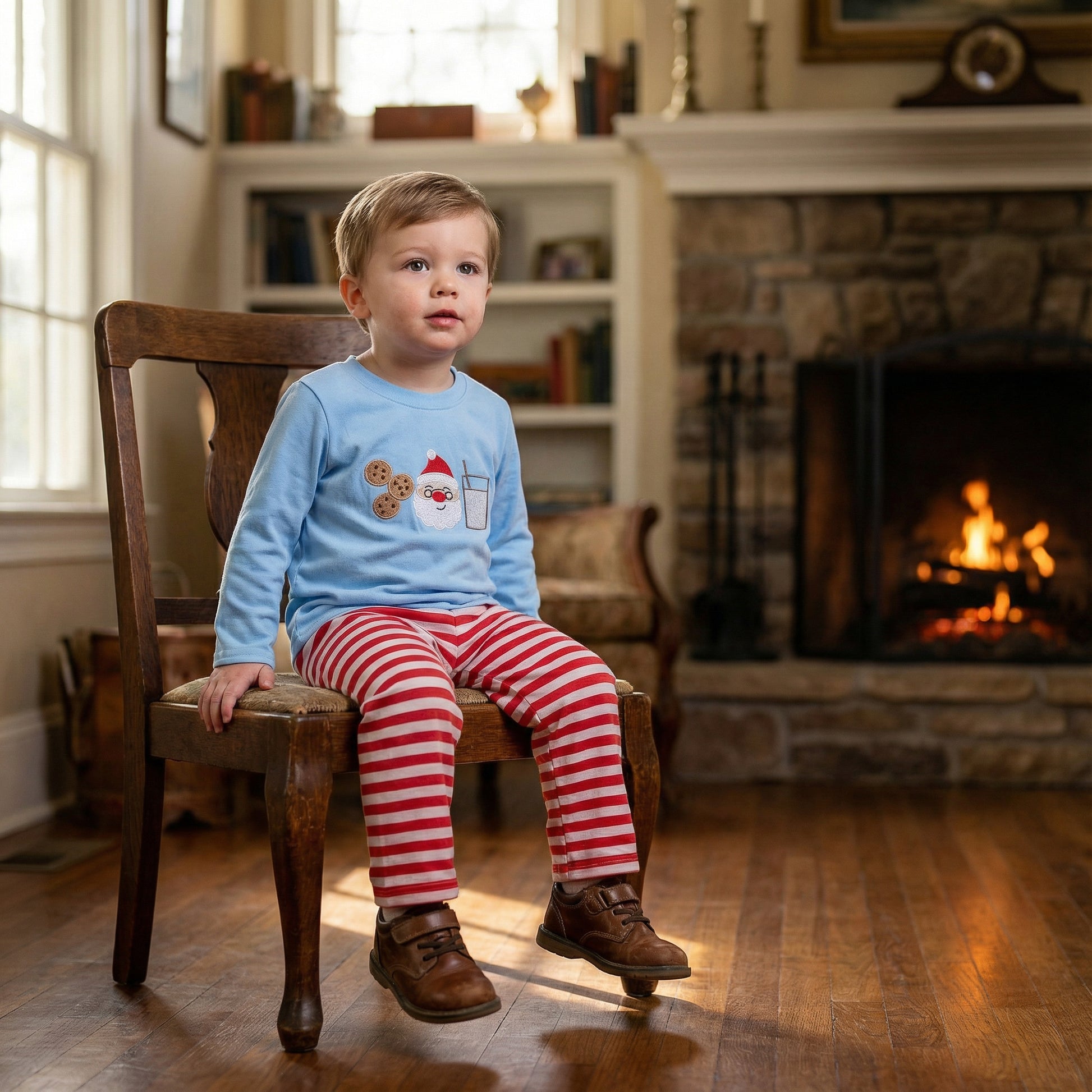 Toddler boy sitting by fireplace wearing blue embroidered Santa milk and cookies shirt and red striped pants.
