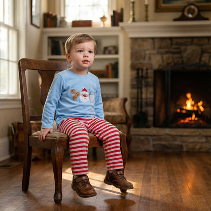 Toddler boy sitting by fireplace wearing blue embroidered Santa milk and cookies shirt and red striped pants.