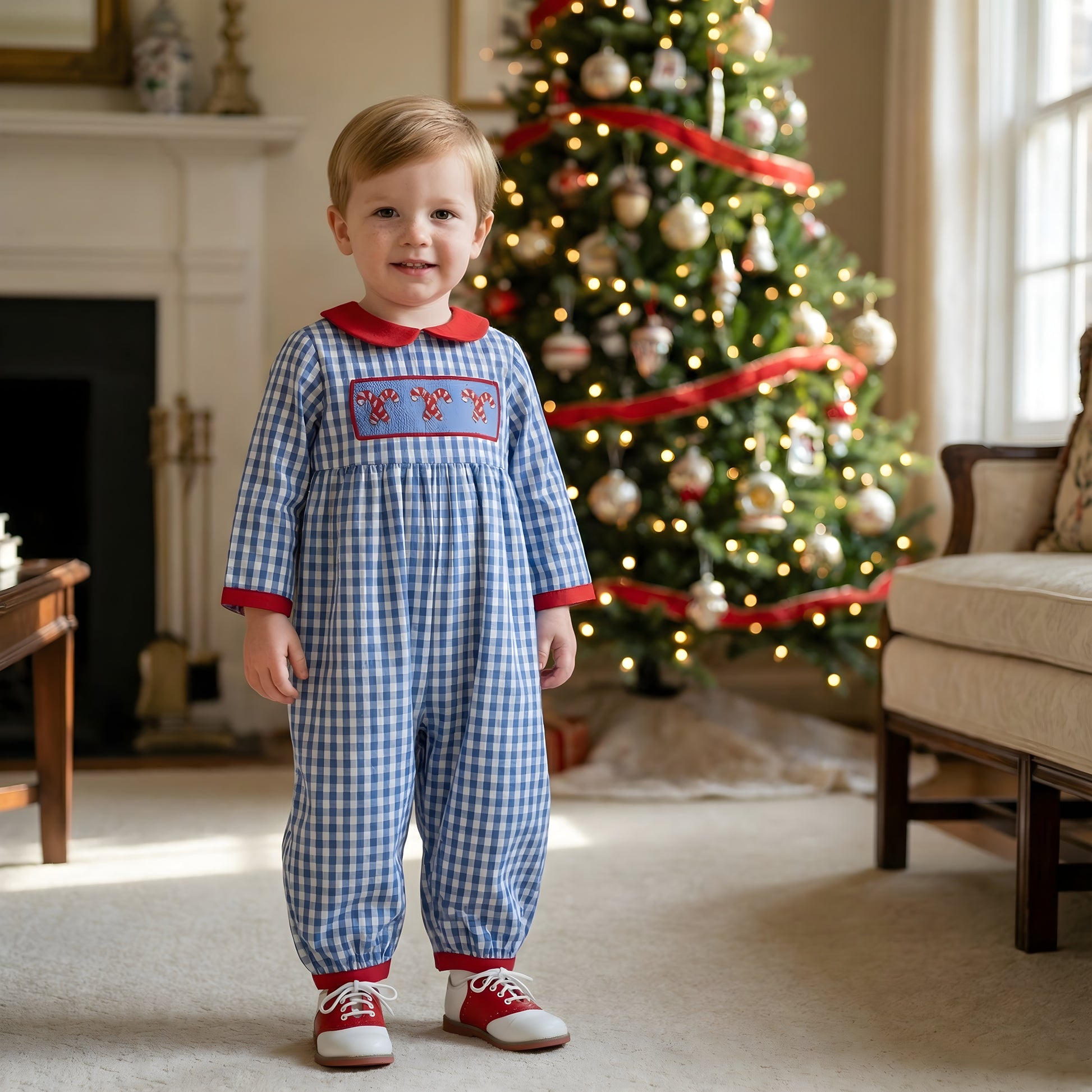 Toddler boy wearing blue gingham hand smocked Christmas candy cane romper standing near holiday tree.