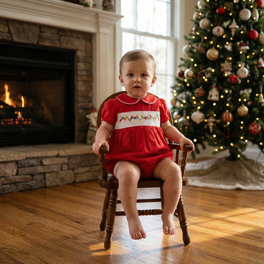 Toddler boy sitting by Christmas tree wearing red smocked bubble with colorful lights embroidery.