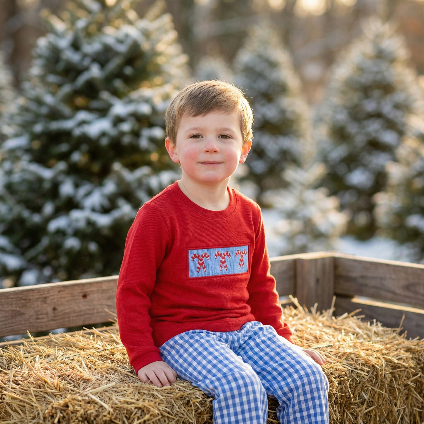 Boy in red smocked candy cane shirt and blue gingham pants sitting on hay bales at a snowy Christmas tree farm.