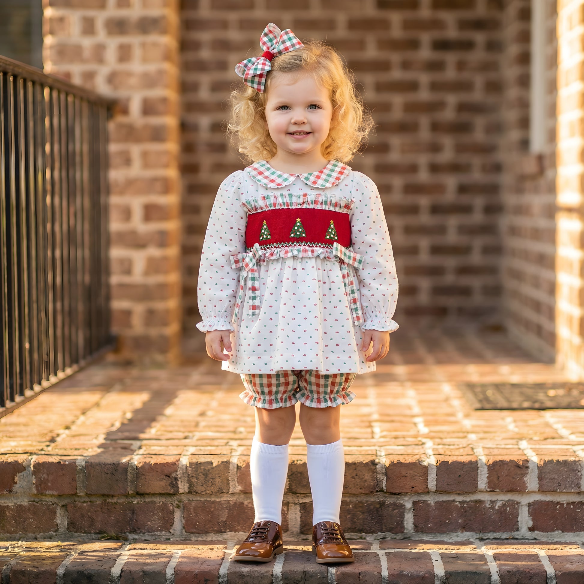 A smiling toddler girl with curly blonde hair stands on brick steps wearing a festive Christmas outfit. She is dressed in a white long-sleeved top featuring a red smocked bodice with embroidered Christmas trees, a plaid Peter Pan collar, and matching plaid ribbons. She wears coordinating plaid bloomers, a large plaid hair bow, white knee-high socks, and brown patent leather shoes. The background shows a blurred brick house exterior and a black railing in warm sunlight.