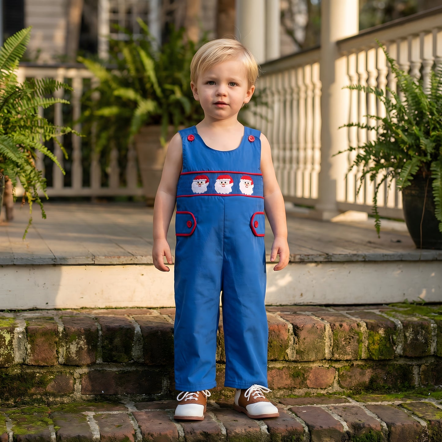 Toddler standing on brick steps wearing a Boys Navy Blue Santa Smocked Christmas Jon Jon featuring red trim and embroidered Santa faces.