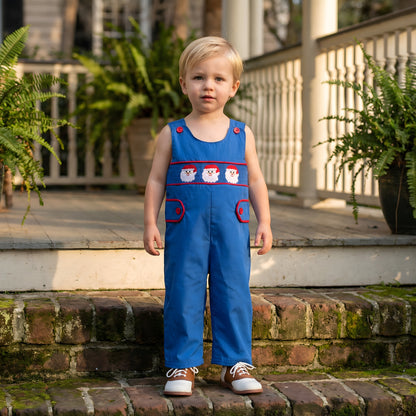 Toddler standing on brick steps wearing a Boys Navy Blue Santa Smocked Christmas Jon Jon featuring red trim and embroidered Santa faces.