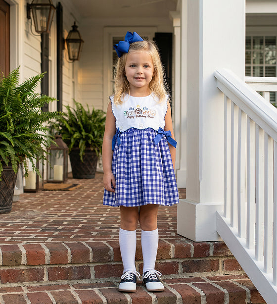 Little girl wearing a blue gingham nativity embroidered dress with side bows standing on brick porch steps.