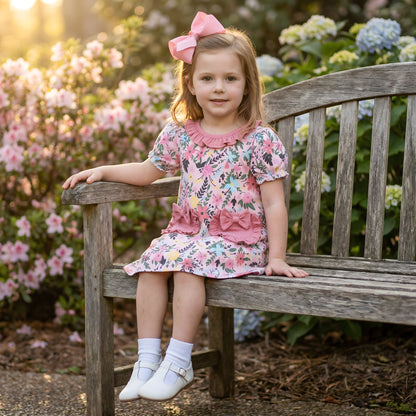 Little girl sitting on a wooden bench wearing the pink floral Field Of Flowers Spring Pocket Dress with ruffle details and a matching hair bow.