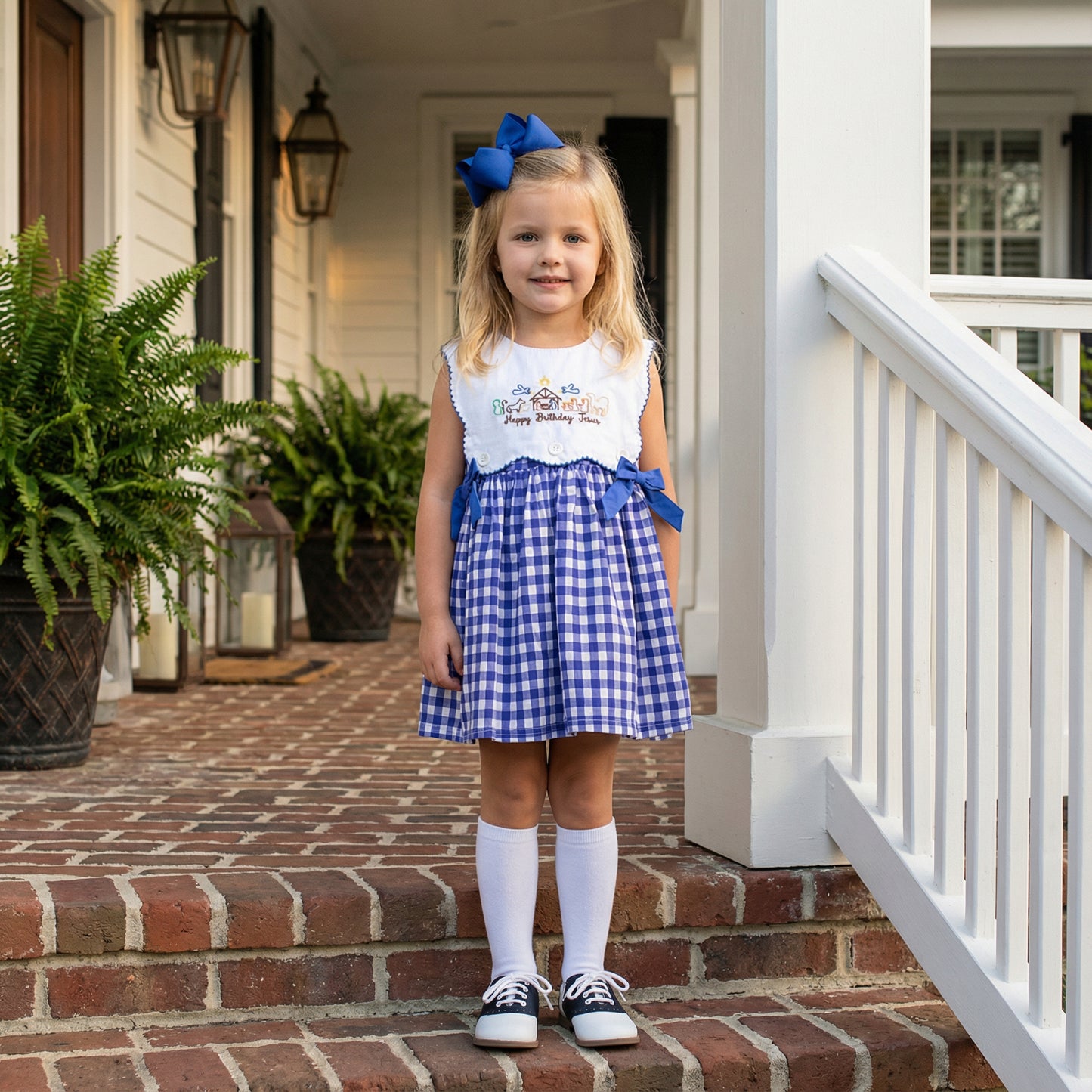 A young girl with blonde hair stands on brick porch steps wearing a blue and white gingham dress. The dress features a white bodice with nativity embroidery and the text 'Happy Birthday Jesus,' accented with blue side bows. She wears a matching large blue hair bow, white knee-high socks, and navy and white saddle shoes. 