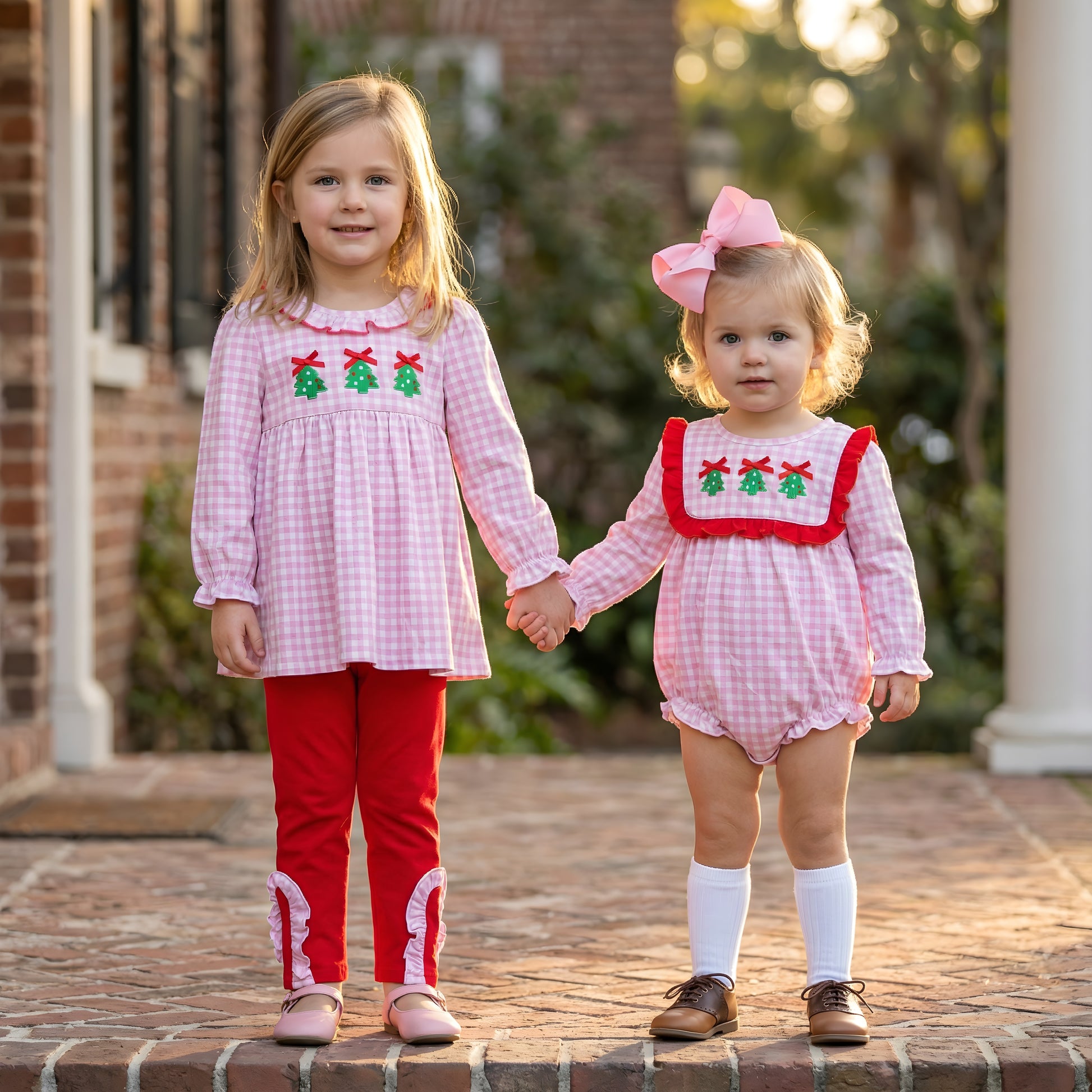 Two young girls, sisters, wearing matching pink gingham Christmas outfits with smocked tops featuring embroidered green Christmas trees and red bows. The older girl wears a tunic top with red ruffled pants, and the younger girl wears a ruffled bubble romper with a large pink bow in her hair. They are holding hands outside on a brick pathway. This children's boutique clothing is perfect for holiday photos.
