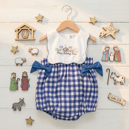 An overhead photograph captures a baby's romper on a wooden hanger, adorned with a "Happy Birthday Jesus" embroidered nativity scene on the white bodice and blue gingham shorts with blue bows. The romper is surrounded by various small wooden nativity figurines, including the holy family, shepherds, kings, animals, a stable, and stars, all arranged on a white wooden plank surface. A wooden scroll with "Luke 2:11" is also present.
