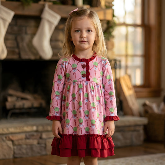 A young girl with blonde hair and a pink bow stands indoors, wearing a pink long-sleeved nightgown with a red ruffled hem and trim. The nightgown has a repeating pattern of green-haired elf figures and candy canes. She is positioned in front of a stone fireplace with stockings hanging from the mantel and a sunlit window to her right.