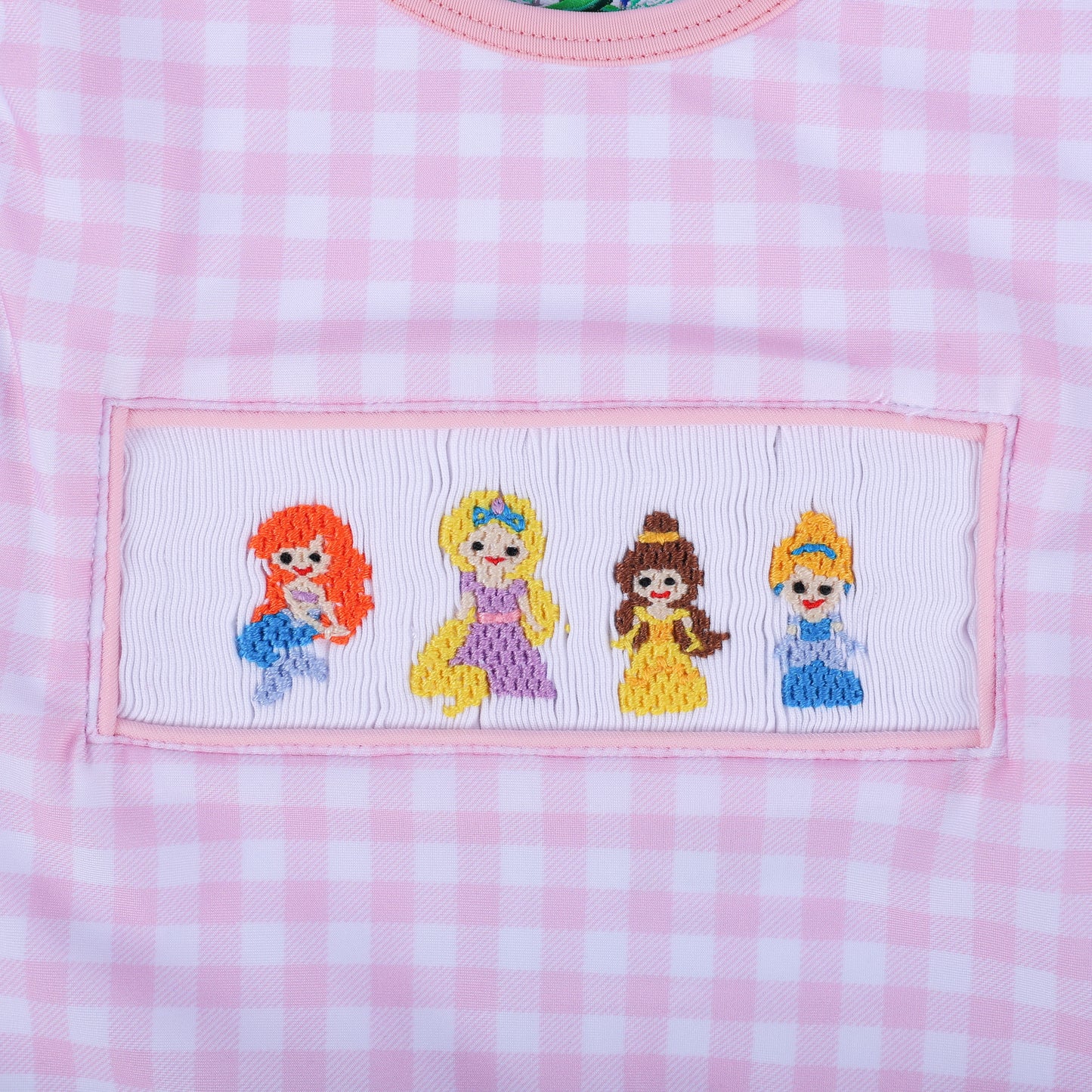 Detailed macro view of the four hand embroidered fairytale princesses on the smocked panel of the gingham swimwear.