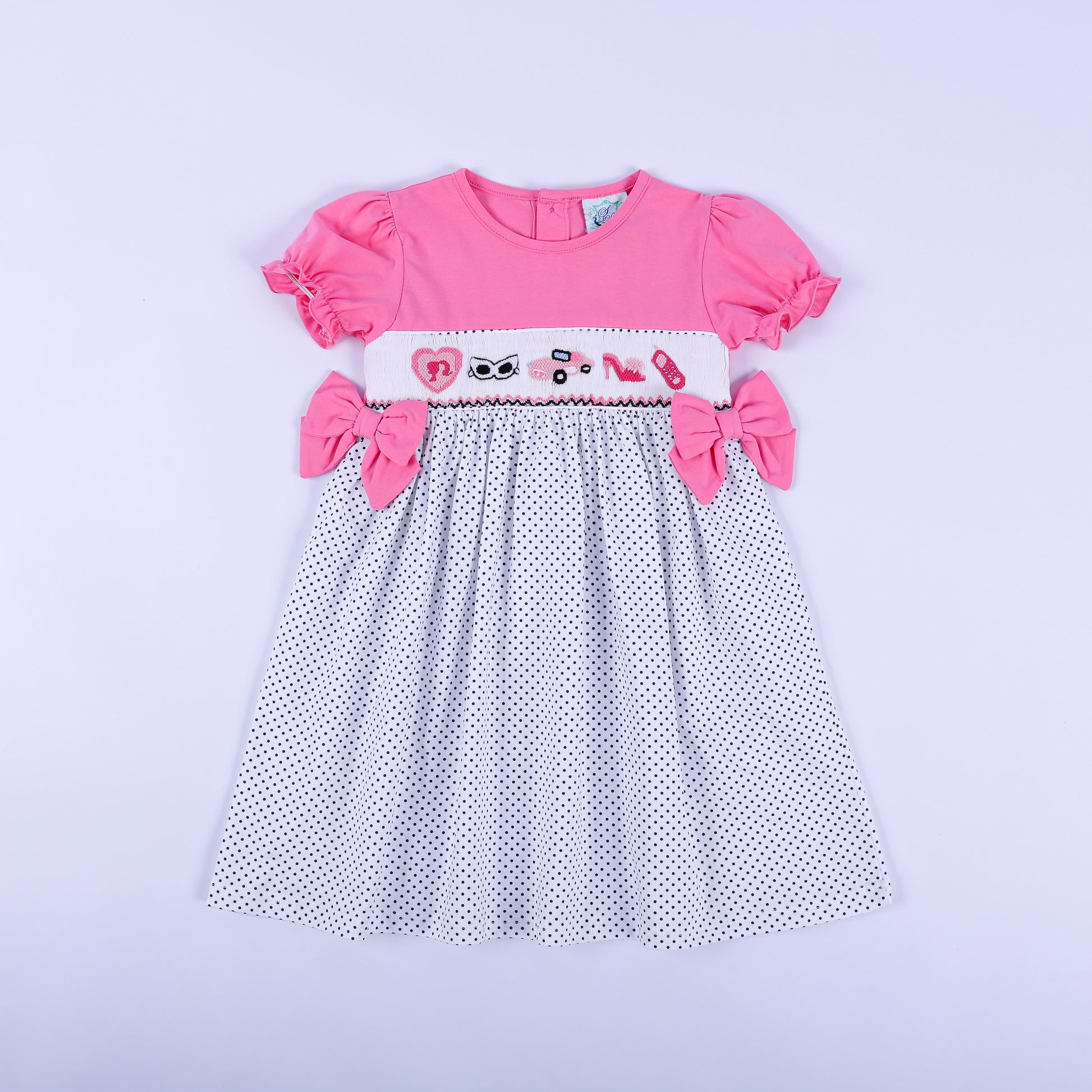 Girls pink polka dot hand smocked dress featuring hand embroidered fashionista motifs and side bows front view.