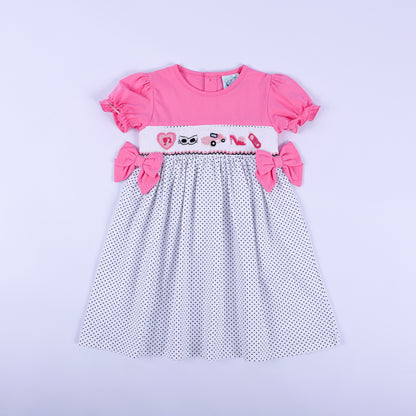 Girls pink polka dot hand smocked dress featuring hand embroidered fashionista motifs and side bows front view.