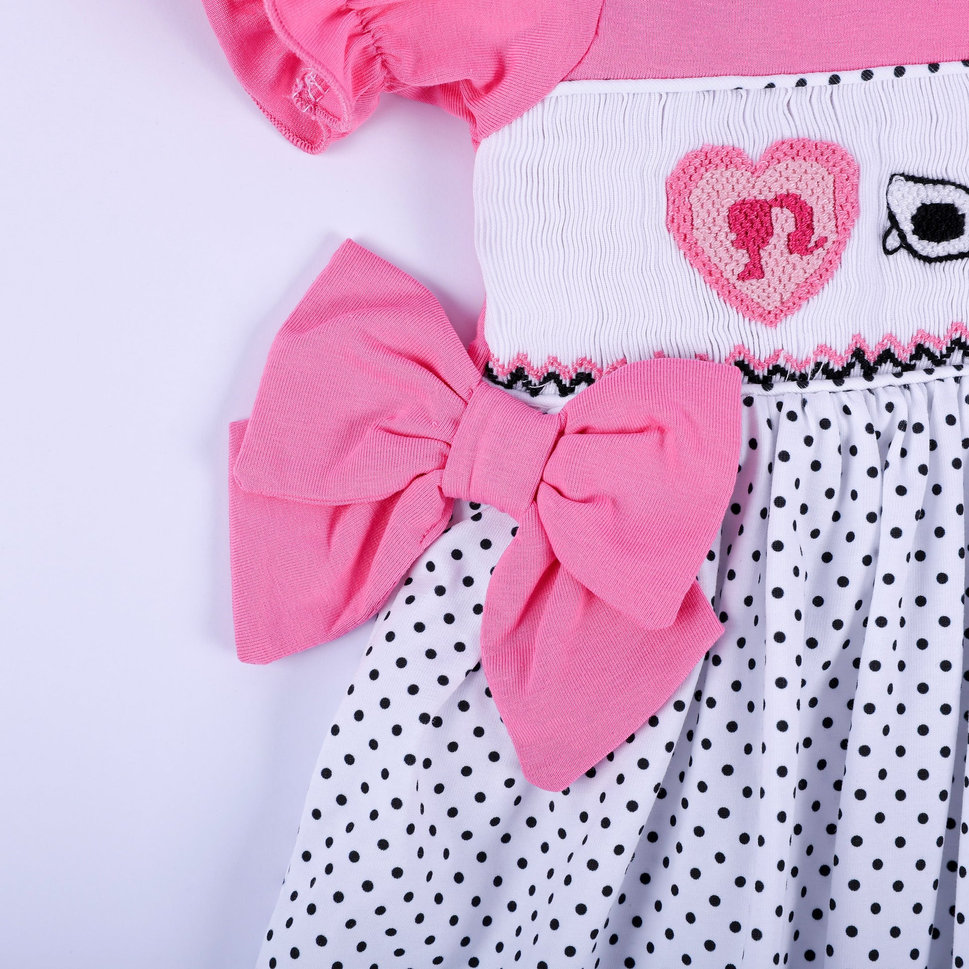 Detail shot of pink side waist bow accent and hand embroidered heart motif on girls polka dot smocked dress.