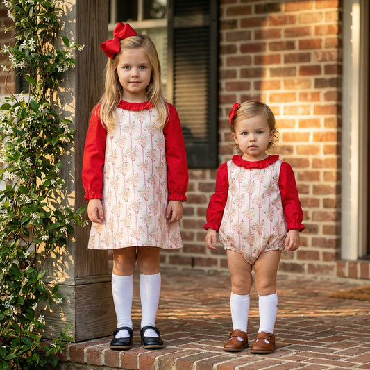 Two children standing on a brick porch wearing matching holiday outfits. Both have long-sleeve red tops with white pinafore-style garments featuring a pink bow and green leaf pattern. The older child wears a knee-length dress with white knee-high socks and dark shoes, while the younger child wears a romper with white knee-high socks and brown shoes. Each has a large red bow accessory in their hair.