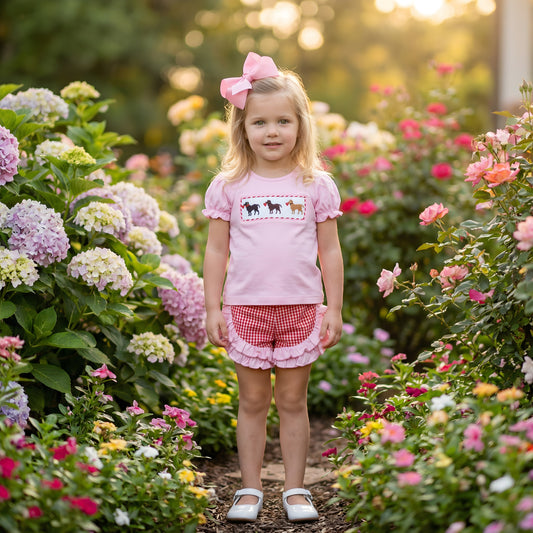 Toddler girl wearing Hand Smocked Valentines Puppy Dogs girls set featuring a pink top with smocked puppies and red gingham ruffle shorts in a garden.