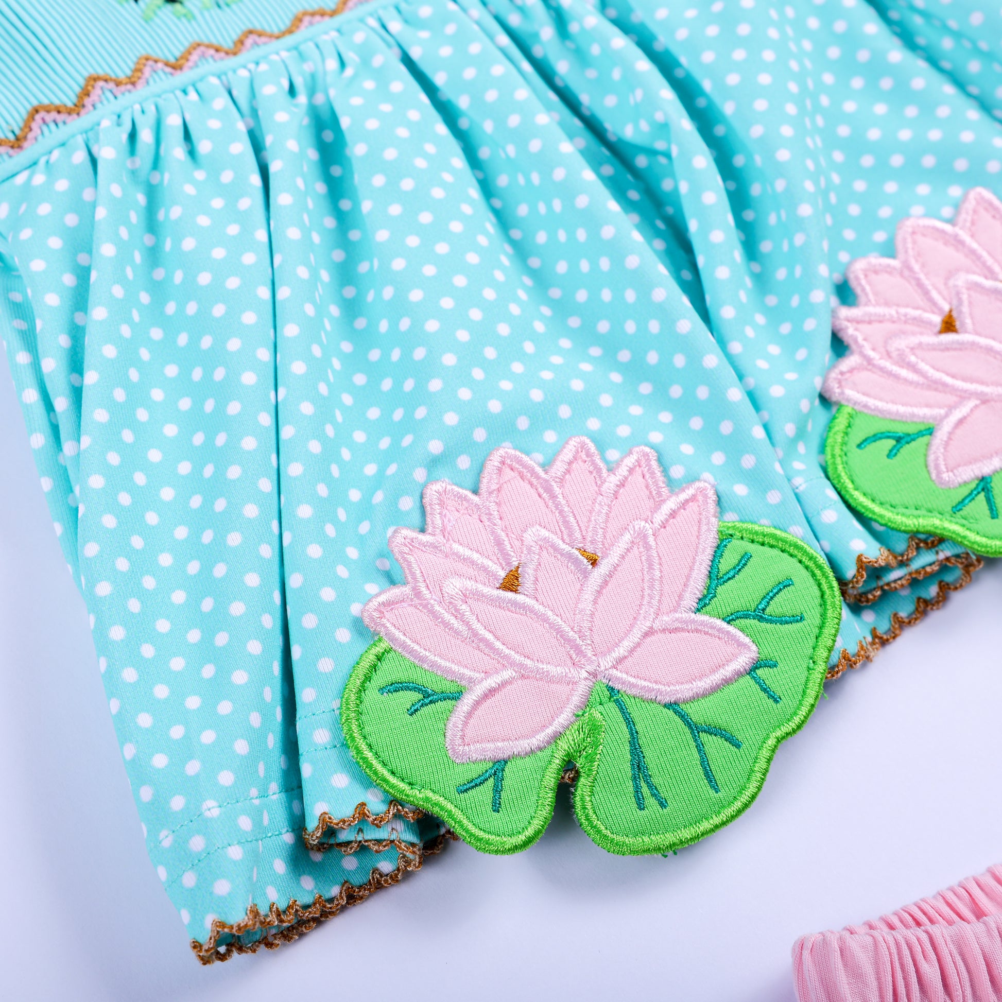 Detailed close-up of pink embroidered lotus flower appliques on the hem of aqua polka dot smocked top.